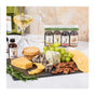 A slate board displays the Cheese Pairing Collection by Runamok: cheese wedges, blue cheese, grapes, crackers, pecans, chocolate, and rosemary with Runamok infused syrups for perfect pairing. White wine and a wine bottle are in the background.