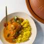 A plate of yellow rice and curry with cilantro garnish sits next to the VitaClay® Chef VITACLAY NourishingPot™ (VM1688-9), a lidded, brown natural clay cooker, on a marble surface.