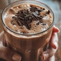 A person holds a glass of Kuli Kuli Foods Organic Moringa Green Smoothie + Protein, frothy and topped with chocolate shavings.