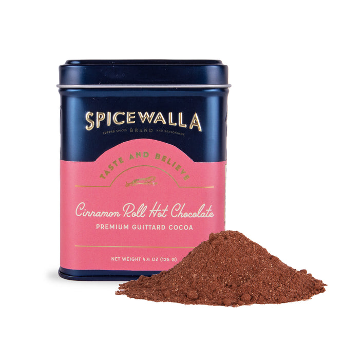 A tin labeled Spicewalla Deluxe Cinnamon Roll Hot Chocolate sits with cocoa powder in front. The dark blue tin features a pink label and gold text, offering a rich, indulgent cinnamon roll and chocolate experience.