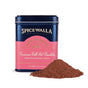A tin labeled Spicewalla Deluxe Cinnamon Roll Hot Chocolate sits with cocoa powder in front. The dark blue tin features a pink label and gold text, offering a rich, indulgent cinnamon roll and chocolate experience.
