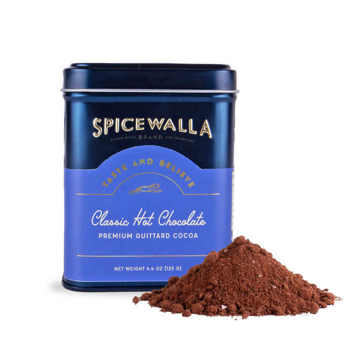 A blue Spicewalla tin labeled Deluxe Classic Hot Chocolate stands next to a small heap of loose, artisanal hot chocolate powder made with rich dark chocolate.