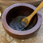 A wooden bowl filled with the dark gray Rose & Hibiscus Clay Face Mask by Chagrin Valley Soap & Salve, made with organic hibiscus and French Pink Kaolin Clay, sits on a stone surface, with a wooden spoon partially buried in the powder.