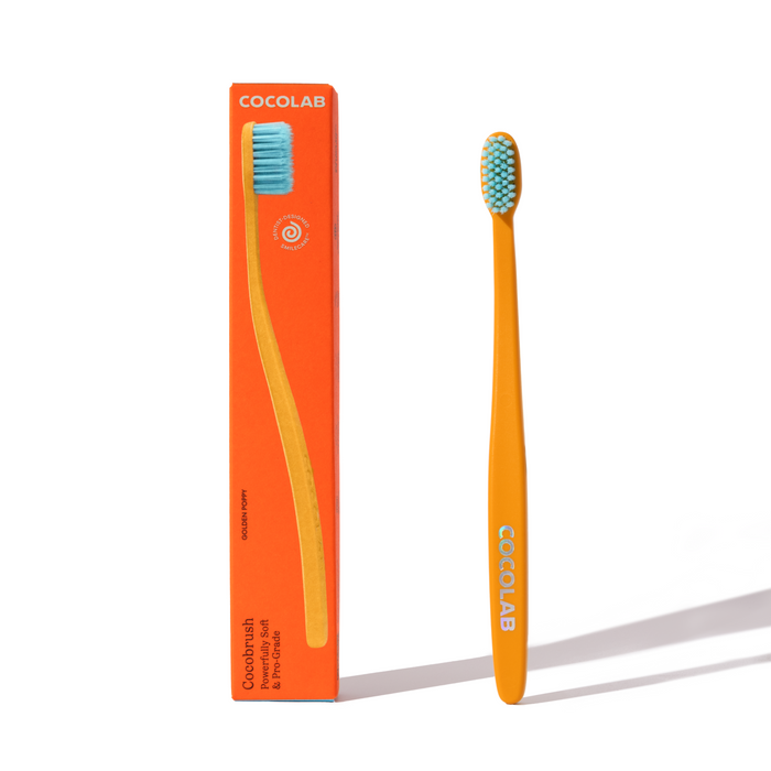 An orange Cocobrush Toothbrush by COCOLAB with blue soft bristles stands upright beside its matching orange box, which displays an illustration of the toothbrush and product information.