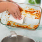 Using the Full Circle Heavy Duty Coconut Scrubber Sponge, a person cleans baked-on residue and orange-stained spots from a glass baking dish while a pot rests in the sink below.