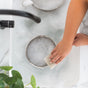 A person cleans a metal pot in a soapy white sink using the Full Circle Heavy Duty Coconut Scrubber Sponge, while a green potted plant rests on the marble countertop nearby.