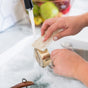 A person uses the Full Circle Heavy Duty Coconut Scrubber Sponge to clean a metal measuring cup with soap suds under running water at the kitchen sink. A fruit basket can be seen in the background.
