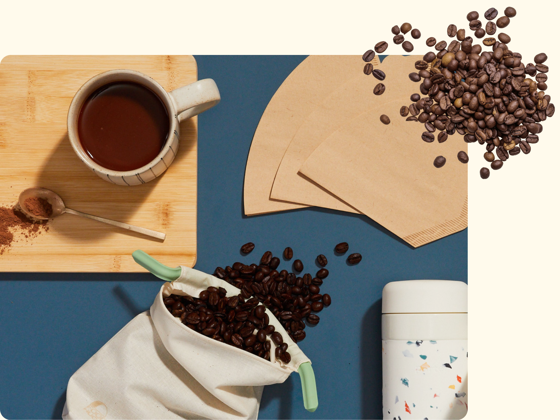 A flat lay of coffee beans, a white mug of coffee on a wooden board, a spoon with coffee grounds, paper filters, a white thermos, and coffee beans spilling from a cloth bag on a blue background.