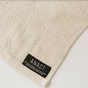A close-up of the Anact Hemp and Organic Cotton Wash Cloth Bath Sheet in beige, showing its textured sustainable fabric and a black label with 