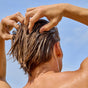 Facing away from the camera against a blue sky, a person with wet, light brown hair massages their scalp to promote scalp health using Phil’s Conditioner - Strengthening + Thickening. Their shoulders and upper back are visible.