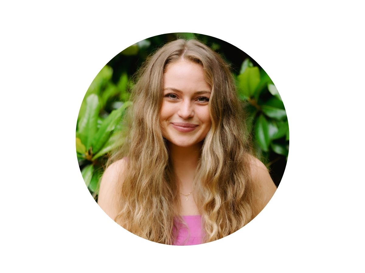 A young woman with long wavy blonde hair, wearing a pink top, smiles in front of green leafy plants. The photo has a circular crop with a white border.