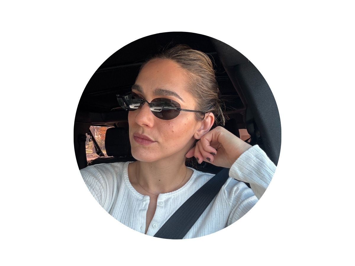 Woman wearing sunglasses and a white top, sitting in a car with a seatbelt on, resting her head on her hand, looking out the window.