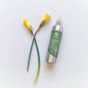 The grab green Room and Fabric Freshener - Vetiver, in a metal spray bottle with a green label, containing essential oils, is beside two closed yellow flower buds on long green stems against a soft white background.