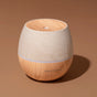 A round Drift Linen Diffuser by Plant Therapy, featuring a wood-grain finish and fabric accent, sits on a brown surface with a small mist hole and a gentle ambient light glowing on top.