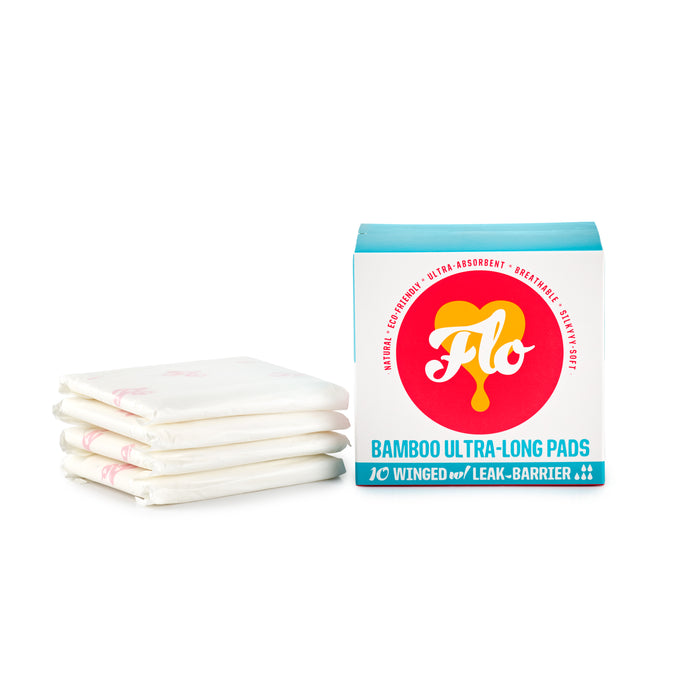 Three individually wrapped sanitary pads are stacked next to a box labeled FLO Bamboo Ultra-Long Pads from Here We Flo, highlighting features such as organic bamboo, super-absorbency, wings, and breathability. The box is vibrantly designed with red and teal text.