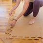 A person kneels on a wooden floor while connecting the puzzle-like pieces of Yoloha Yoga Cork Floor Mat by hand. The non-toxic, interlocking mats are being assembled, though the person’s face is not visible.