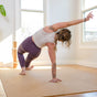 Wearing workout clothes, a person practices a yoga pose on one hand atop the Yoloha Yoga Cork Floor Mat in a sunlit room with large windows, enjoying the comfort of sustainable flooring and the peace of practicing on a non-toxic surface.