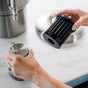 A person’s hands replace a Rorra Countertop Filter, a cylindrical black water filter from Rorra, and a metal cap on a stainless steel countertop next to a metal appliance.