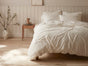 A cozy bedroom with a Coyuchi Classic Organic Sateen Duvet Set in white, a wooden nightstand holding a vase and jug, light wood floors, a ceramic jug with branches, and light paneled walls adorned with framed art.