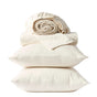 Two cream-colored pillows stacked with a folded flat sheet and rolled-up fitted sheet from the Coyuchi Classic Organic Sateen Sheet Set, all GOTS certified organic cotton, neatly arranged against a white background.