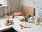 A marble kitchen counter displays covered bowls, a wooden cutting board, Coyuchi Conserve Organic Produce Bags with onions and greens, fabric napkins, and dried orange slices—all illuminated by natural light in a zero waste setup.
