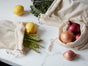 Three Conserve Organic Produce Bags by Coyuchi, made from organic cotton, sit on marble—one holds lemons and asparagus, another holds onions, and leafy greens fill the third—a perfect zero-waste shopping swap.