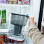 A person places a large Epic Water Filters USA Nano XP Water Filter Pitcher, designed to reduce bacteria and PFAS, inside a refrigerator next to milk, soda cans, and other groceries.