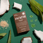 A Jackfir The Bushwhacker Bar Soap box rests on a green surface, surrounded by coconut pieces, green leaves, clear soap drops, and beige granules.