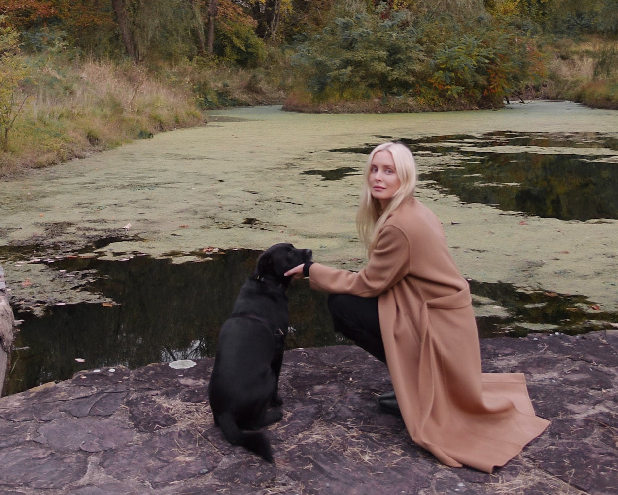 A person in a long tan coat kneels on a stone surface beside a black dog, near a pond covered with green algae, surrounded by autumn trees and foliage.