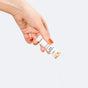 A hand with orange nail polish holds a small white tube labeled Thorne, dispensing Complete Biotic Powder Packets, 3-in-1 Formula as a fine probiotic powder against a plain light background.