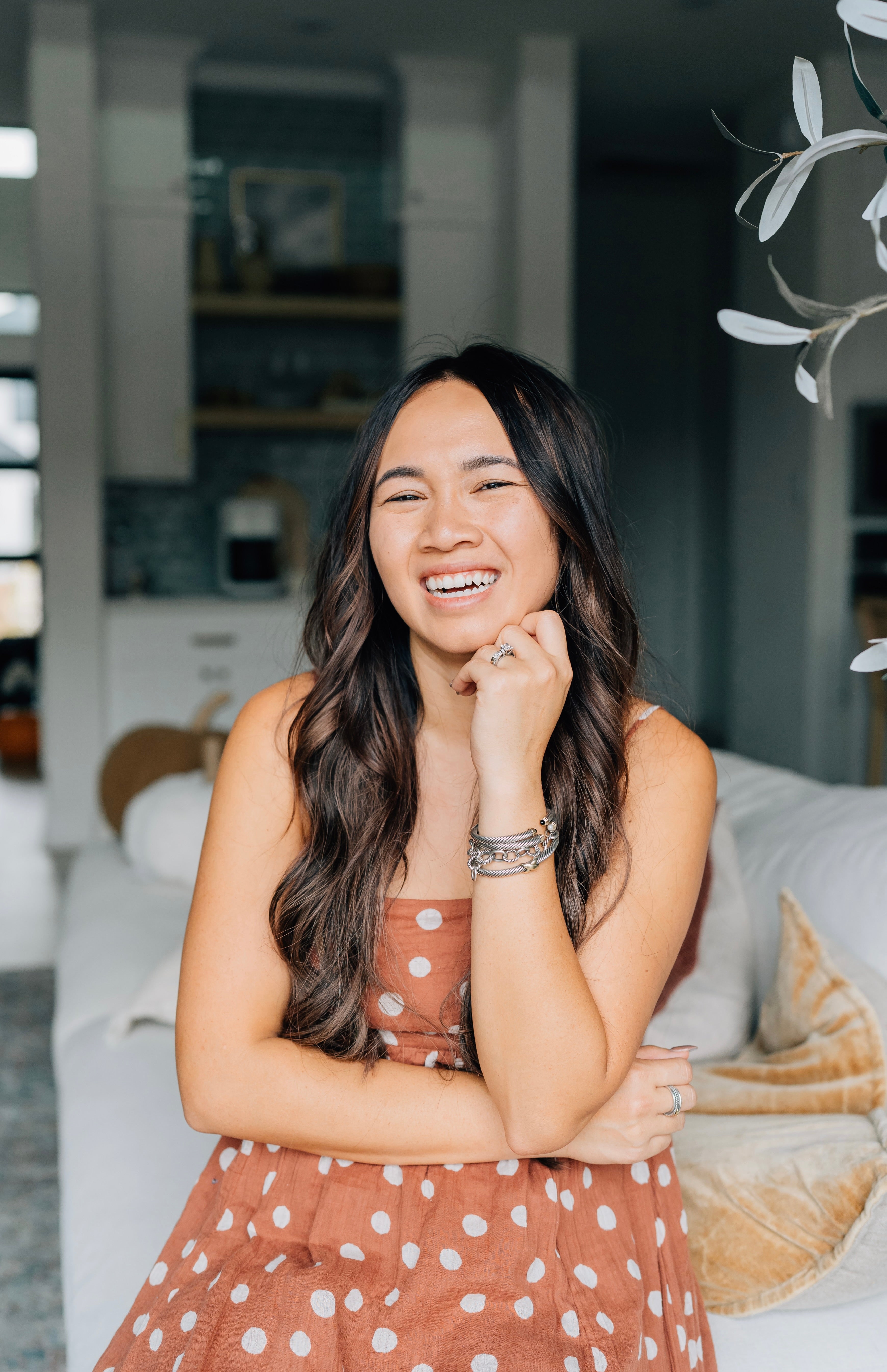 A woman with long brown hair wearing a sleeveless, brown polka dot dress sits on a white couch, smiling and resting her hand on her chin in a bright, modern living room.