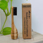 The Me Mother Earth All-in-One Bamboo Travel Toothbrush 2.0 Head - Refill features antimicrobial black bristles and stands upright beside its brown box, with a green plant in the background.
