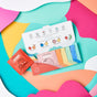 A colorful flat lay shows the Dabble & Dollop Just a Dabble Minis Set, with fun fruit character illustrations. The open box reveals individually wrapped 3-in-1 Bubble Bath packets on abstract pink, teal, and orange paper.