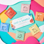 Flat lay of the Dabble & Dollop Just a Dabble Minis Set: six colorful bath sampler packets and a rectangular box on a wavy pink, yellow, blue, and orange background. Each packet displays cute fruit or honey art with natural ingredients.