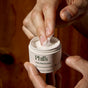 A person scoops Phil's Daily Facial Moisturizer, featuring hyaluronic acid and prebiotics, from a white jar set against a brown background.