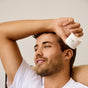 A man with short brown hair and a trimmed beard, wearing a white shirt, smiles while holding a jar of Phil's Daily Facial Moisturizer above his head against a neutral background.