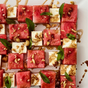 A close-up of a salad featuring watermelon and mozzarella cubes, garnished with mint and nuts, artfully arranged on a white plate, and drizzled with Kosterina Dark Chocolate Balsamic Vinegar for a savory-sweet twist.