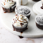 Kosterina Dark Chocolate Balsamic Vinegar is drizzled over chocolate cupcakes with white frosting, some dusted with powdered sugar, and arranged on a marble platter for an irresistible sweet and savory treat.