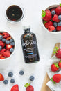A bottle of Kosterina Dark Chocolate Balsamic Vinegar sits on a white surface, surrounded by bowls of fresh strawberries, blueberries, a small bowl of balsamic vinegar, and some loose berries with a towel nearby.