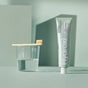 A bamboo toothbrush rests on a clear glass of water beside a tube of Davids Hydroxi™ Premium toothpaste - Whitening + Enamel Health with nano-hydroxyapatite, set against a pale green background with soft lighting and gentle shadows.