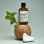 A bottle of Davids Hydroxi™ Oil Pulling Oral Rinse with Nano Hydroxyapatite stands on a whole coconut, accompanied by a mint sprig and a coconut piece in front, set against a pale green background—emphasizing its enamel-supporting formula and long-lasting fresh breath benefits.