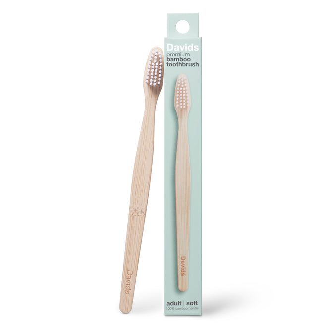 Two Davids Premium Bamboo Toothbrushes - Adult Soft are shown, featuring white soft bristles and "Davids" printed on the handles. One toothbrush stands upright while the other lies beside a light green packaging box labeled Davids Premium Bamboo Toothbrush - Adult Soft.