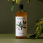 A bottle of Koala Eco Dish Soap Refill with a botanical label rests on a green surface, surrounded by green leaves and yellow flower sprigs, set against a muted green background.