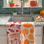The Neighborgoods Dish Towel Set of 2 – Fall Feast features a mushroom “Funging Awesome” towel and a pumpkin towel, both hanging over a sink above veggies, with green tiles and plants in the background.