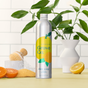 A metal bottle of Grove Co. Dish Soap Refill, adorned with a citrus design and reading dish soap yuzu & mandarin, sits on a yellow surface. Sustainable and plant-based, the scene includes halved citrus fruits, a white vase with green leaves, and a scrub brush against a white tile background.