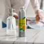 A countertop scene features a silver bottle of Grove Co. Dish Soap Refill with a lemon design. A clear soap dispenser complements it, while in the background, someone in a light sweater efficiently cleans a dish using plant-based ingredients and a brush.