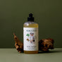 A bottle of Koala Eco Dog Wash with a black cap sits between driftwood on a green surface and background. The label displays a dog and plant graphics, emphasizing quality coat care.