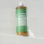A bottle of Dr. Bronners Pure-Castile Liquid Soap rests on a tiled surface with soap suds at its base and a white tiled wall in the background, known for its vegan formula and gentle cleansing.