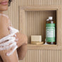 A person with soapy arms stands by a wooden shower shelf, holding a bar of All-One! soap and a large bottle of Dr. Bronner’s Pure-Castile Liquid Soap in almond scent, set against vertical wood paneling.