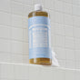 A 32 fl oz (946 ml) bottle of Dr. Bronners Pure-Castile Liquid Soap sits on a white tiled shower ledge, its blue and white label marking this vegan, gentle cleanser. A bit of foam rests at the base of the Baby Unscented variety.